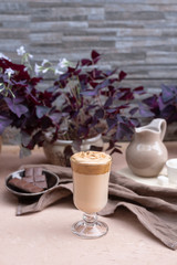 A popular Korean drink during self-isolation is Dalgona coffee. A delicious modern cold beverage with chocolate. Creamer, napkin is on the table. On a background is potted plants. Vertical still life.