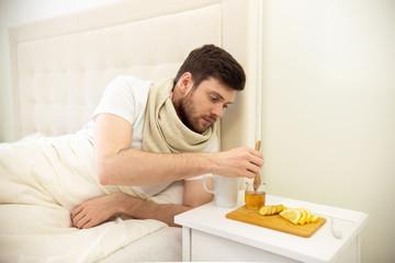 Sick Man in Bed Adding Honey into Tee. Man Ill in Bed Self Healing. Self Treatment at Home. Man Drinking Hot Tea in Bed.