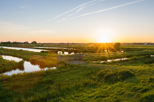 Sun sets over the wide open Dutch polder landscape. Green meadows intersected with ditches filled with water.