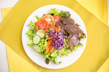 vegetable salad top with grilled sliced beef in white plate served dressing in a dipping bowl on a yellow placemat