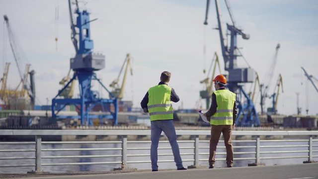 Wide shot of two shipbuilding engineers walking at dockside, examining vessels under construction and discussing plans