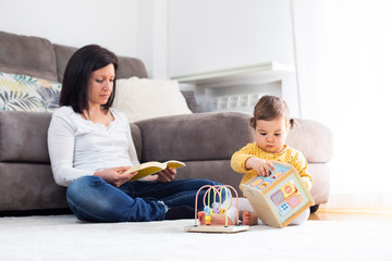 Madre leyendo un libro en casa mientras su hija pequeña que juega con un juguete educativo de madera a su lado