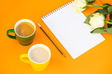 Two cup of cappuccino, paper notepad with pencil and white roses on the orange background. Empty space for text.