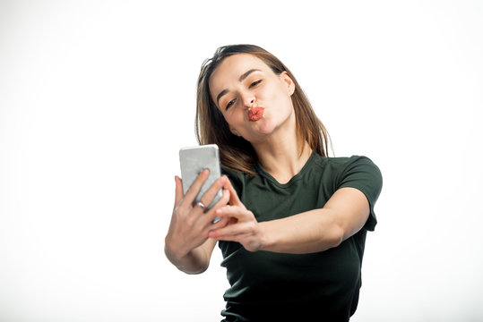 Pretty girl makes her lips duck and take a self portrait with her smart phone. Cute lovely young woman making duck face and taking selfie with mobile phone over white background.