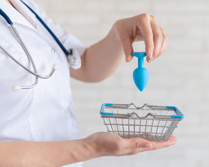 Faceless gynecologist puts anal plug in miniature shopping baskets. The doctor holds the device for...