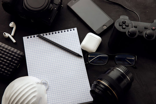 Hipster flatlay with camera, lens, glasses, joystick, wireless headphones, notebooks, pen, respirator on a black table. Top view of the workspace. Creative desktop for photographer, traveler, blogger