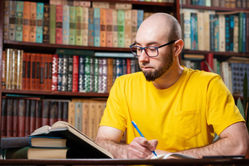 A bald, bearded man with glasses is writing something in a notebook, looking at a book. There are...