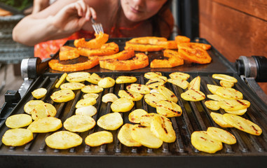 Potato chips and butternut squash pieces grilled on electric grill, focus on bright potatoes circles, blurred young woman in background
