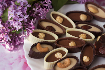 Bouquet of lilac flowers and chocolate candies with filling and nuts,  chocolates with nuts on a plate, background. Sweets made of black, milk and white chocolate with hazelnuts, almonds and cashews