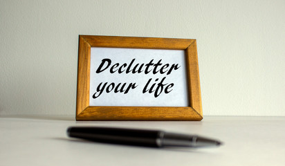 Wooden picture frame with inscription 'declutter your life' on beautiful white fon. Pen on the table.