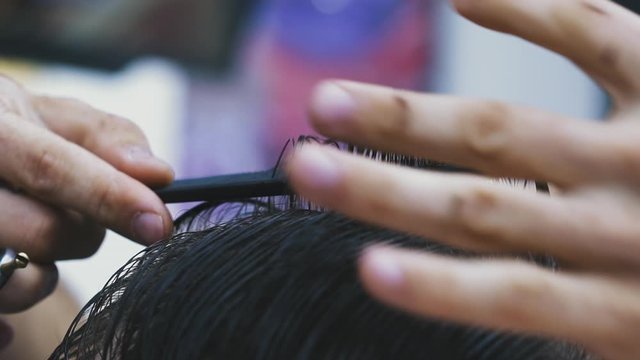 CU, slow motion: young barber does stylish haircut to client with black hair using scissors and comb in modern brightly lit beauty salon close view