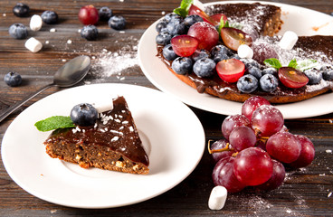 piece of chocolate cake with fresh berries, blueberries, grapes, powdered sugar ,  mint on a white plate on a wooden