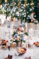 Wedding dinner table reception. Bouquet in a vase on a table of pink and white roses and blue hydrangea, with the number of the table on the plate. Burning candles in high glass candlesticks.