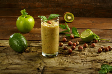 A glass of green smoothie on a wooden table near avocado apple lime nuts.