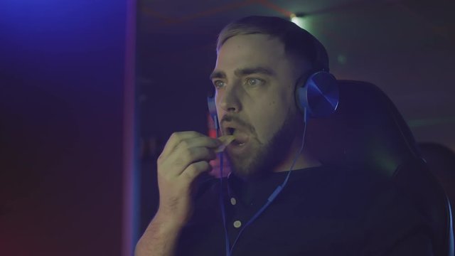 Medium shot of bearded Caucasian young man wearing earphones sitting in front of computer screen, playing online game and eating potato chips