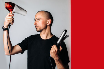 a bald brutal man, holding a hair dryer and Curling tongs in his hand, dries his hair and baldness .