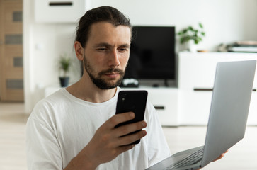 Man with beard, working at home with a laptop in front of him and speaking with a friends or business partners or clients with a smartphone and sitting on a floor. Freelancer working from home.
