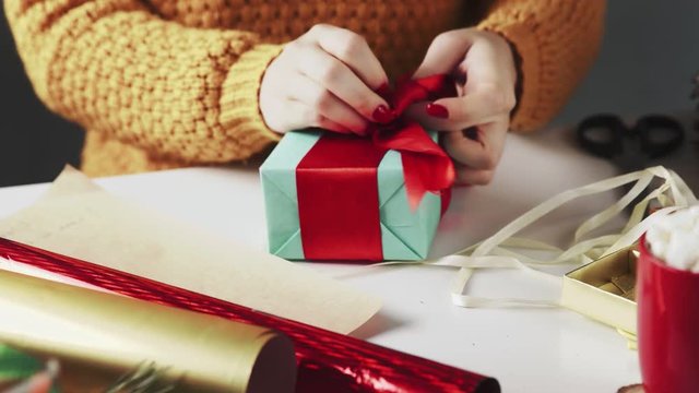Female hands wrapping Christmas gift in blue paper and tying red ribbon