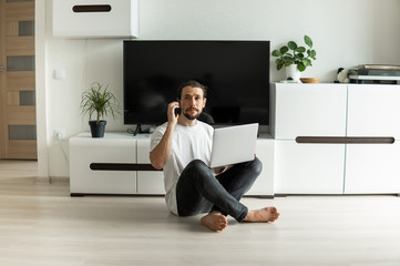 Man with beard, working at home with a laptop in front of him and speaking with a friends or business partners or clients with a smartphone and sitting on a floor. Freelancer working from home.