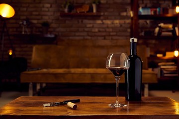 Wine glass and a bottle of red wine on table at home, cosy dark living room in background. Autumn mood with dark and warm colors.