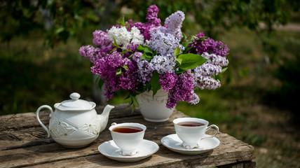 Lilac spring flowers in vase on wooden background. Tea coffee cup on table during breakfast time
