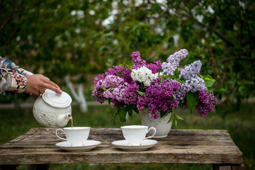 Lilac spring flowers in vase on wooden background. Tea coffee cup on table during breakfast time