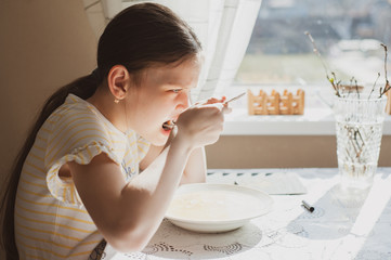 Teen girl eats a spoon of soup at a table at home in the kitchen