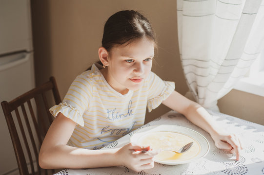 Teen Girl Eats A Spoon Of Soup At A Table At Home In The Kitchen