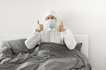 Portrait of man in protective white suit and face mask sitting in his bed in his bedroom because of covid-2019 and wait for a end of coronavirus quarantine. Self isolation.