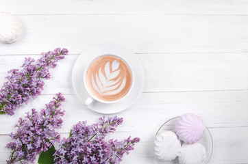 Lilac, coffee with latte art and marshmallow on white wooden table. Romantic morning. Flat lay