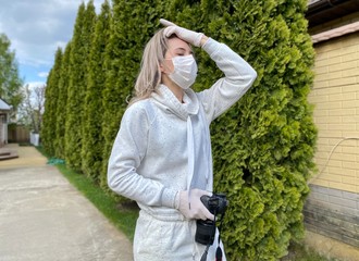 beautiful young woman on the street in a protective flu mask looks away, holds a dsl camera in her hands on a background of fir