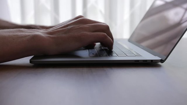A man puts a laptop computer on a table in front of a window with white curtains and starts typing, then removes the laptop and leaves.