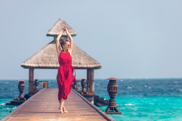 Joyful young happy girl in red jumpsuit standing on a wooden pier near water bungalows and enjoying ocean, summer breeze and sound of the waves during vacation. Happy holiday travel concept.