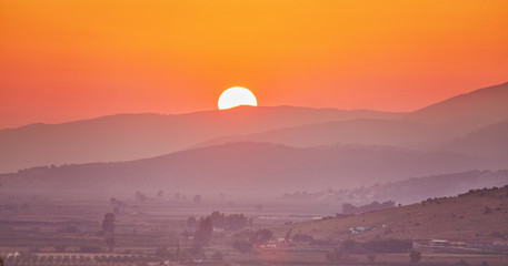 Panorama of beautiful mountains covered with trees at dawn in summer. Bright large orange sun disc....