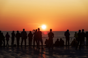 Fototapeta na wymiar Silhouettes of people against the setting sun on deck of a cruise ship. Calm ocean and clear orange sky in evening time. Cruise vacations background