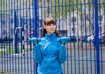 A beautiful young girl in a tracksuit doing bicep exercises with dumbbells in the fresh air.