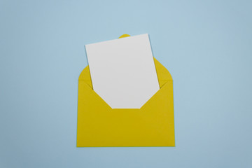 Minimalistic composition with a beautiful festive yellow mustard envelope, white blank card on a trendy blue pastel background. Mockup with envelope and blank card. Place for text. Flat lay. Top view
