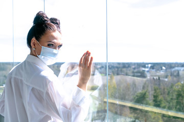 Fototapeta premium In a medical mask looks out the window. Concept: self-isolation, virus, coronavirus. be at home