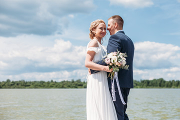 Panoramic view at young couple bride in white and groom in blue standing among lake, posing and embracing. Conceptual wedding at nature outdoor in summer season. Wedding, family, love story concept.
