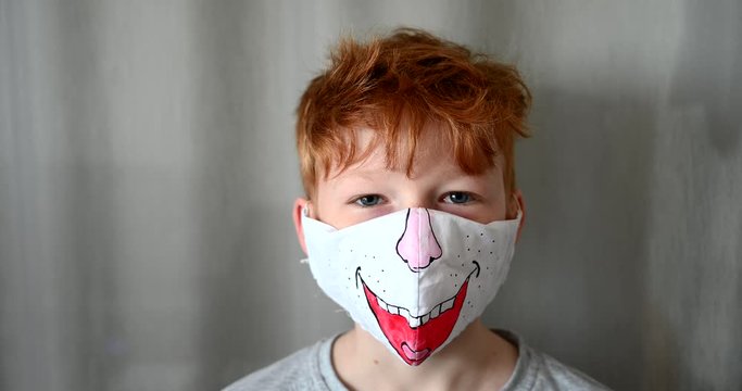 Little reddish red hai boy looking into camera wearing original face mask painted during coronavirus pandemic. 