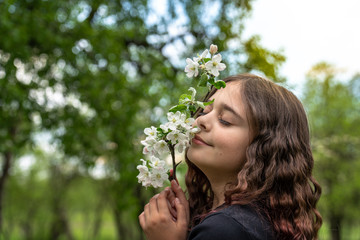 Obraz premium Tender curly brunette girl with closed eyes holds blooming branch near her face, blurred background