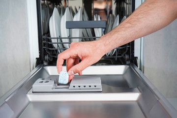 Putting tab into dishwasher, close up. A man hand holding dishwasher detergent tablet. Dishwasher machine full loaded.