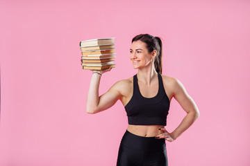 Beautiful, attractive, muscular girl holding heavy set of books on one hand, and with her second hand showing her large muscles. Demonstrating pumped up body in photo studio on pink background