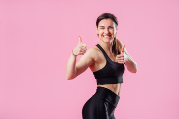 Portrait of an excited young brunette sportive muscular girl showing two thumbs up isolated over pink background. Attractive girl is happy with her figure. Ready for the summer season