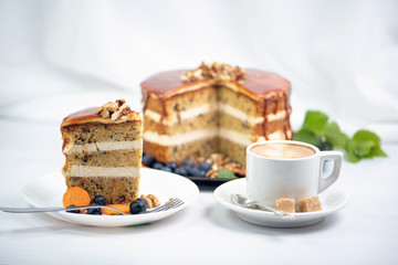 a cup of coffee with two pieces of sugar on a saucer and next to it a piece of carrot cake on a white plate, covered with caramel and decorated with nuts on top, in the background is a cake and a spri