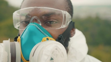 African scientist in white chemical suit protection wearing glasses respirator full face mask in prevention of coronavirus infection. Outdoor portrait. Close-up. - Powered by Adobe