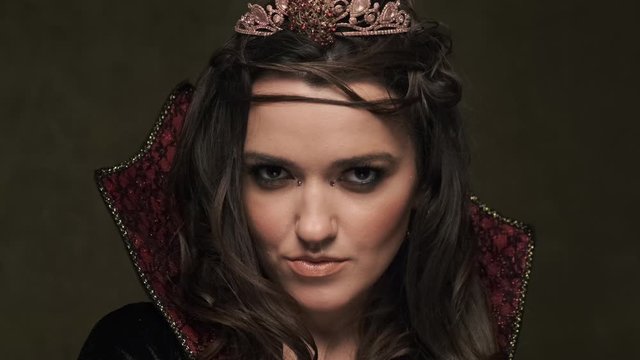 Beautiful queen at dark background. Female in cloak and crown. Portrait of vampiress looking at camera. Close-up shot in 4K, UHD