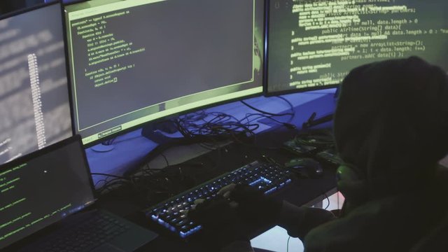 High angle view of unrecognizable person wearing hoody and headphones on his neck sitting at desk at multiple computers and coding