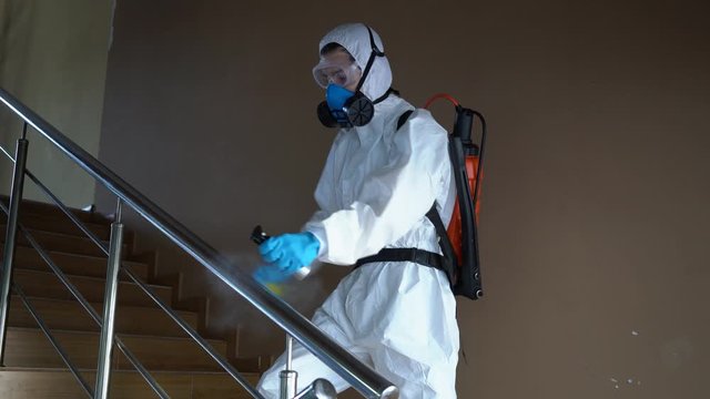 Sanitizer disinfects railing touch surfaces on coronavirus covid-19 quarantine with antibacterial sprayer. Worker in gas mask and hazmat protective suit cleans handrails in city public place.