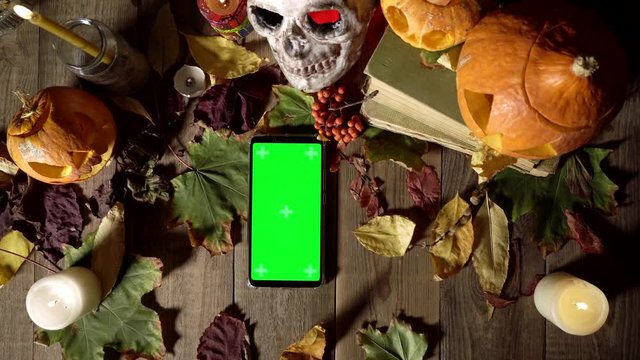 Top view of smart phone laying at the autumn composition of leaves. Hallowene theme with skull, leaves and candles at the wooden table. Hands put the phone down then taking it away.